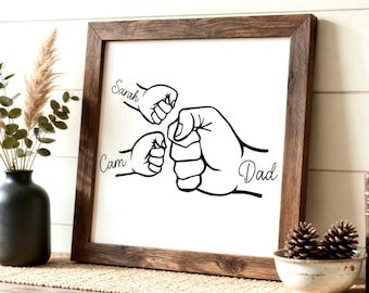 May include: A framed wall art print featuring a black and white illustration of three fists bumping. The names "Sarah", "Cam", and "Dad" are written above or near each fist. The frame is made of dark brown wood.