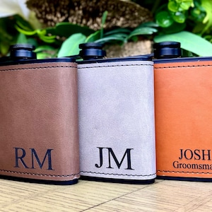 May include: Three leather-wrapped hip flasks with personalized engravings. The flasks are brown, gray, and orange. The engravings read "RM", "JM", and "JOSH Groomman".