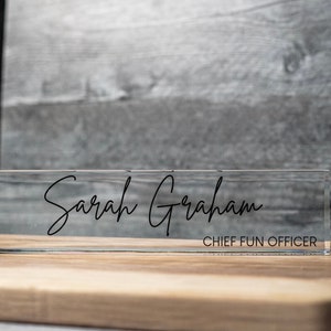 May include: A clear acrylic desk nameplate displaying the name "Sarah Graham" in black script. Beneath the name, the title "CHIEF FUN OFFICER" is printed in a smaller font. The nameplate is positioned on a wooden surface.