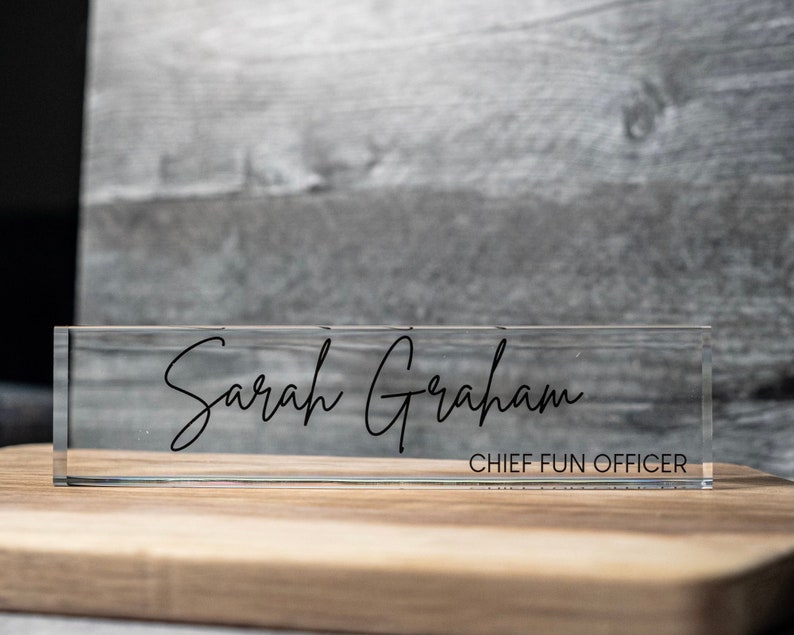 May include: A clear acrylic desk nameplate displaying the name "Sarah Graham" in black script. Beneath the name, the title "CHIEF FUN OFFICER" is printed in a smaller font. The nameplate is positioned on a wooden surface.