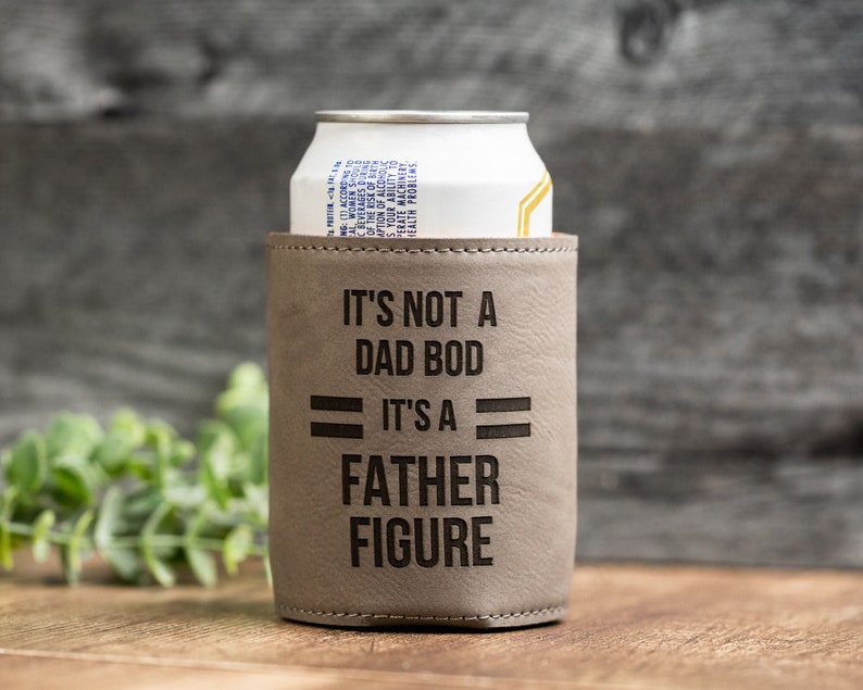 May include: A gray leatherette can cooler with the text "IT'S NOT A DAD BOD IT'S A FATHER FIGURE" in black. A white and yellow can is inside the cooler. The background is a wooden surface.