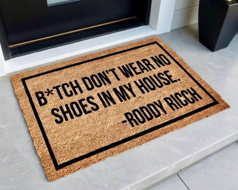 May include: A rectangular doormat made of natural coir fiber. The mat has a black border and features the text "B*TCH DON'T WEAR NO SHOES IN MY HOUSE. -RODDY RICCH" in bold black letters. The mat is placed in front of a door.