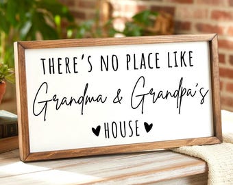 May include: A framed wooden sign with the text "THERE'S NO PLACE LIKE Grandma & Grandpa's HOUSE" in black script on a white background. The frame is a warm brown color, and two black hearts are below the word "HOUSE".