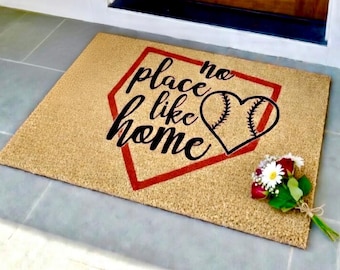 May include: A rectangular doormat with the phrase "no place like home" in black script, inside a red baseball diamond outline. A black heart-shaped baseball is on the right. A small bouquet of flowers rests on the mat.