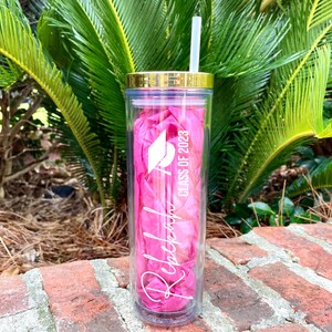Puede incluir: Un vaso rosa y transparente con tapa dorada y una pajita blanca. El vaso tiene el texto "Rebecca" y "Class of 2023" con un gr&aacute;fico de gorro de graduaci&oacute;n.
