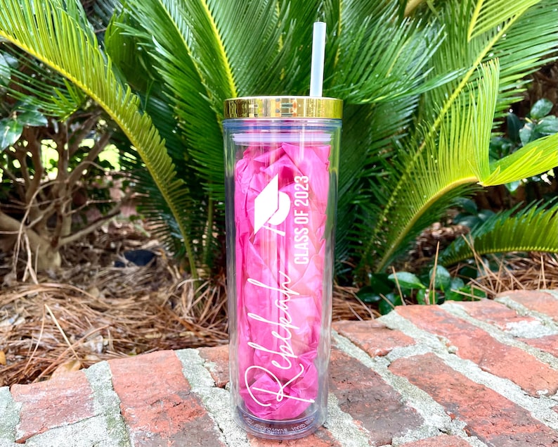 Puede incluir: Un vaso rosa y transparente con tapa dorada y una pajita blanca. El vaso tiene el texto "Rebecca" y "Class of 2023" con un gr&aacute;fico de gorro de graduaci&oacute;n.