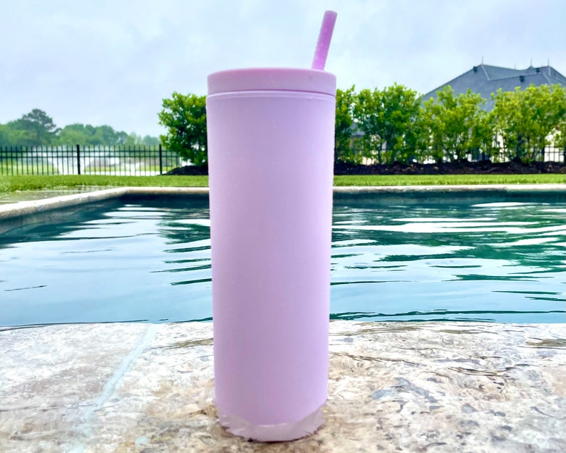 May include: A light pink stainless steel tumbler with a lid and a pink straw. The tumbler is sitting on a stone surface next to a swimming pool.