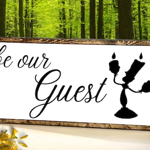 Puede incluir: Un letrero de madera con fondo blanco y texto negro que dice "be our guest". Una silueta negra de Lumiere, el candelabro de La Bella y la Bestia, est&aacute; en el lado derecho del letrero.