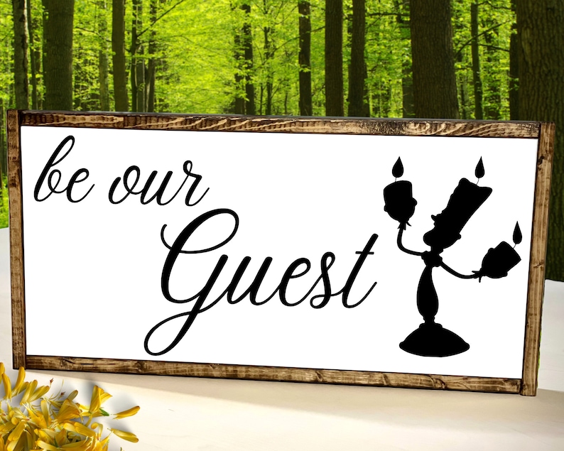 Puede incluir: Un letrero de madera con fondo blanco y texto negro que dice "be our guest". Una silueta negra de Lumiere, el candelabro de La Bella y la Bestia, est&aacute; en el lado derecho del letrero.
