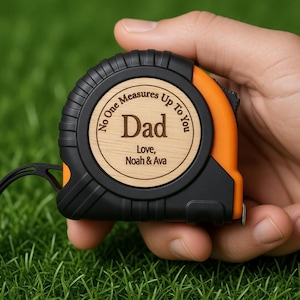 May include: A black and orange tape measure with a wooden center. The wooden center has the word "Dad" engraved, along with the text "No One Measures Up To You" and the names "Love, Noah & Ava".