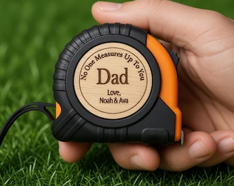 Puede incluir: Un flexómetro negro y naranja con un centro de madera. El centro de madera tiene la palabra "Dad" grabada, junto con el texto "No One Measures Up To You" y los nombres "Love, Noah & Ava".