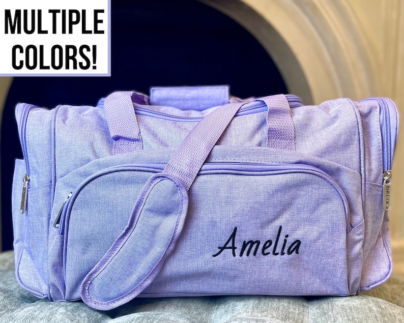 May include: A lavender-colored duffel bag with a zipper closure and a black embroidered name "Amelia".
