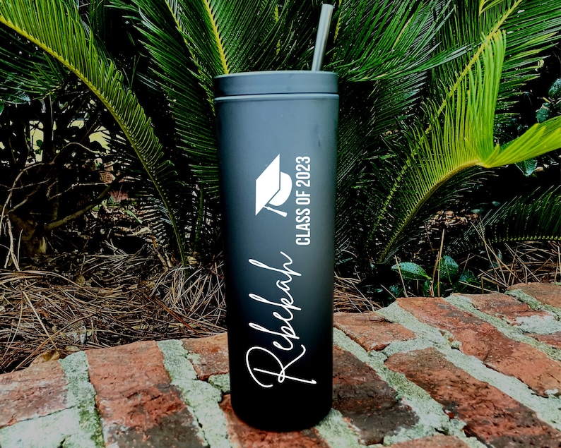 Puede incluir: Vaso de acero inoxidable negro con un gorro de graduaci&oacute;n blanco y el texto "Class of 2023" y el nombre "Rebekah" en una fuente cursiva.