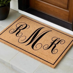 May include: A rectangular doormat with the letters "RMK" in a black, elegant script. The mat is a natural tan color with a black border, placed in front of a wooden door.