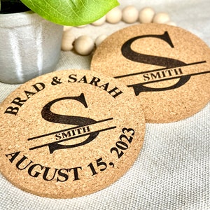 May include: Two round cork coasters with a personalized design. Each coaster features the letter "S" with the last name "SMITH" underneath. One coaster has the text "BRAD & SARAH" and "AUGUST 15, 2023" around the letter. The coasters are light brown.