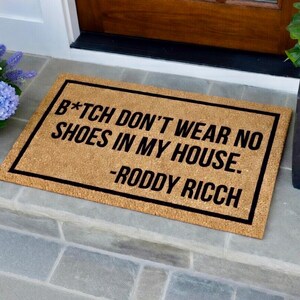 May include: Rectangular doormat made of natural coir fiber with a black border. The mat features the text "B*TCH DON'T WEAR NO SHOES IN MY HOUSE. -RODDY RICCH" in black, perfect for a front door.