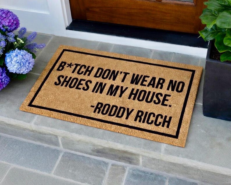 May include: Rectangular doormat made of natural coir fiber with a black border. The mat features the text "B*TCH DON'T WEAR NO SHOES IN MY HOUSE. -RODDY RICCH" in black, perfect for a front door.