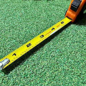May include: A yellow measuring tape with black markings and a black and silver metal hook. The tape is extended to show the numbers 1, 2, 3, 4, and 5. The tape is lying on a green surface.