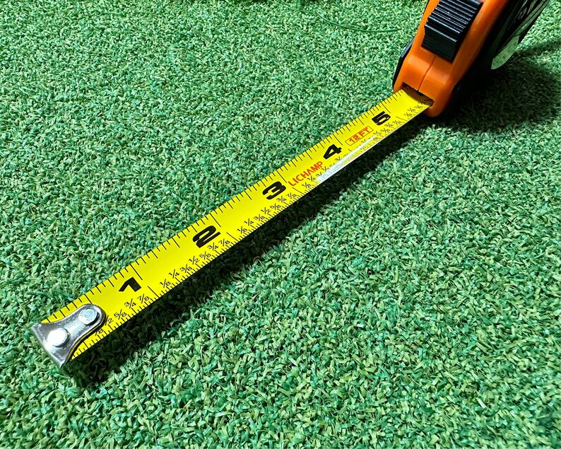 May include: A yellow measuring tape with black markings and a black and silver metal hook. The tape is extended to show the numbers 1, 2, 3, 4, and 5. The tape is lying on a green surface.
