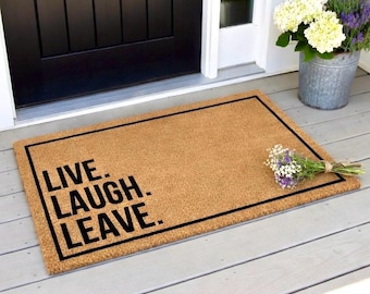 May include: A rectangular doormat made of natural brown coir fiber. The mat has a black border and the words "LIVE. LAUGH. LEAVE." in bold black letters. A small bouquet of flowers rests on the mat.