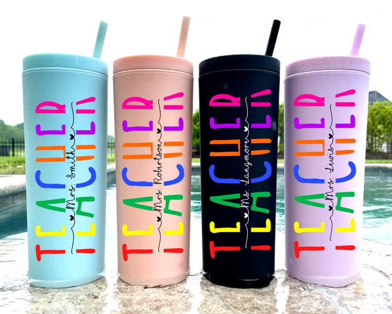 May include: Four colourful stainless steel tumblers with straws. Each tumbler has the word "TEACHER" written in rainbow colours with a teacher's name underneath. The tumblers are in blue, pink, black, and purple.