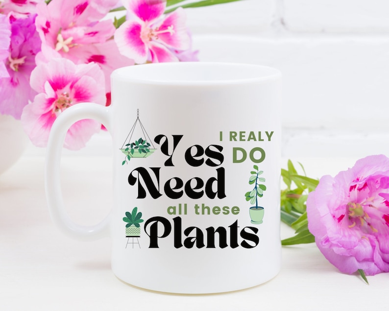 K&ouml;nnte beinhalten: Eine wei&szlig;e Keramiktasse mit einer schwarzen und gr&uuml;nen Grafik, auf der "Ja, ich brauche wirklich all diese Pflanzen" steht. Die Tasse hat einen schwarzen Henkel und steht auf einer wei&szlig;en Oberfl&auml;che.