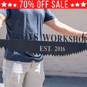 Puede incluir: Un letrero de metal negro en forma de hoja de sierra con el texto "Dad's Workshop Est. 2016" en letras blancas. El letrero est&aacute; siendo sostenido por una persona.