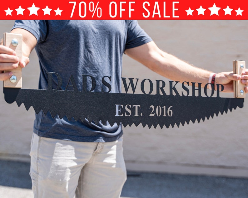 Puede incluir: Un letrero de metal negro en forma de hoja de sierra con el texto "Dad's Workshop Est. 2016" en letras blancas. El letrero est&aacute; siendo sostenido por una persona.