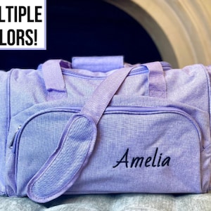 May include: A lavender-colored duffel bag with a zipper closure and a shoulder strap. The bag has a small pocket on the front and the name "Amelia" is embroidered on the front.