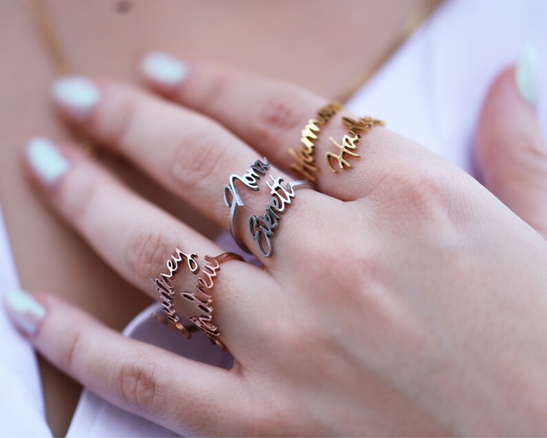 May include: Three gold rings with names engraved on them. The rings are worn on a person's finger. The names on the rings are "Hannah Hall", "Mama Bennett", and "Matthew Andrew".