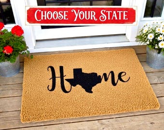 May include: A rectangular doormat with the word "Home" in black script, with the shape of the state of Texas replacing the "O". The mat is tan and sits in front of a white door. A red sign above the door reads "Choose Your State".