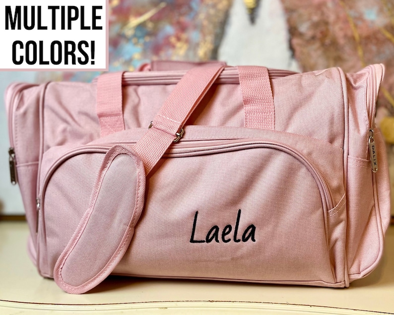 May include: A pink duffel bag with a black embroidered name "Laela" and a matching pink zipper pull. The bag has a shoulder strap and a small zippered pocket on the side.