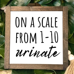 May include: A white wooden sign with brown wood frame, featuring the text "On a scale from 1-10 urinate".