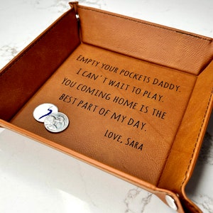 May include: A brown leather valet tray with the text "EMPTY YOUR POCKETS DADDY. I CAN'T WAIT TO PLAY. YOU COMING HOME IS THE BEST PART OF MY DAY. LOVE, SARA" engraved on the inside.