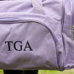 Pu&ograve; includere: Borsa da viaggio in tela di colore viola con una cerniera e un monogramma ricamato nero "TGA" sul davanti.