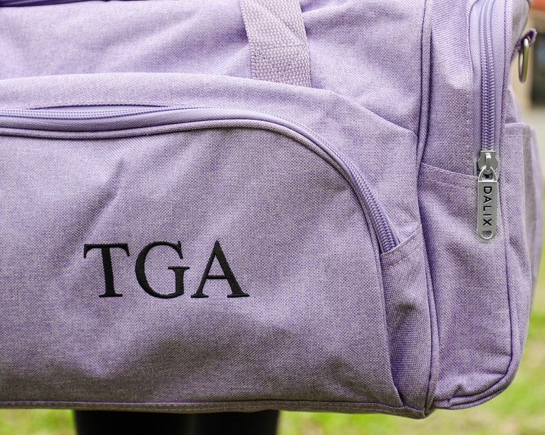 Pu&ograve; includere: Borsa da viaggio in tela di colore viola con una cerniera e un monogramma ricamato nero "TGA" sul davanti.