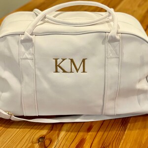 May include: White canvas duffel bag with a gold zipper and a gold embroidered monogram "KM".