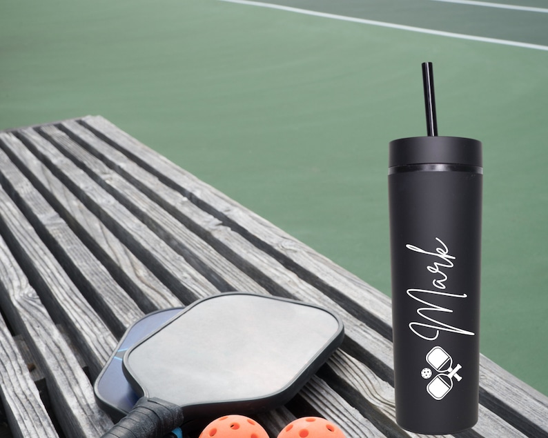 K&ouml;nnte beinhalten: Schwarzer und wei&szlig;er personalisierter Becher mit Strohhalm, mit einem Pickleball-Schl&auml;ger- und -Ball-Motiv und dem Namen "Mark". Der Becher steht auf einer Holzbank mit zwei Pickleball-Schl&auml;gern und zwei orangefarbenen Pickleballs.