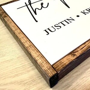 Puede incluir: Un letrero de madera con fondo blanco y marco marr&oacute;n oscuro. El letrero dice "The Justin & Kristi Est." en letras negras.