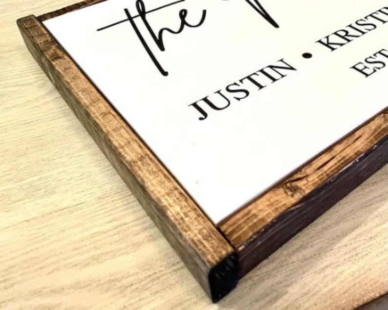 Puede incluir: Un letrero de madera con fondo blanco y marco marr&oacute;n oscuro. El letrero dice "The Justin & Kristi Est." en letras negras.