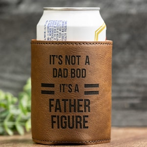May include: A brown leatherette can cooler with the text "IT'S NOT A DAD BOD IT'S A FATHER FIGURE" in black. A white and gold can is inside the cooler. The cooler is designed to keep beverages cold.