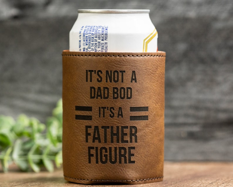 May include: A brown leatherette can cooler with the text "IT'S NOT A DAD BOD IT'S A FATHER FIGURE" in black. A white and gold can is inside the cooler. The cooler is designed to keep beverages cold.