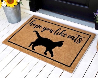 May include: A rectangular doormat made of natural coir fiber. The mat features a black border and the phrase "hope you like cats" in a handwritten style font. A black silhouette of a cat is in the center.