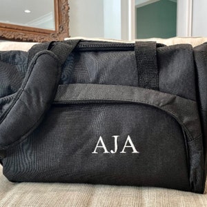 May include: Black travel duffel bag with a white embroidered monogram "AJA".