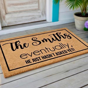 May include: A rectangular doormat with a natural coir fiber surface. The mat features a black border and the words "The Smiths eventually, HE JUST HASN'T ASKED YET!" in a black, cursive font. The mat is placed on a wooden porch.