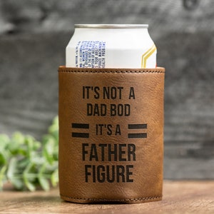 May include: A brown leatherette can cooler with the text "IT'S NOT A DAD BOD IT'S A FATHER FIGURE" in black. A white and silver can is inside the cooler. The background is a wooden surface.