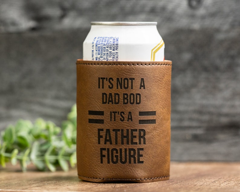May include: A brown leatherette can cooler with the text "IT'S NOT A DAD BOD IT'S A FATHER FIGURE" in black. A white and silver can is inside the cooler. The background is a wooden surface.