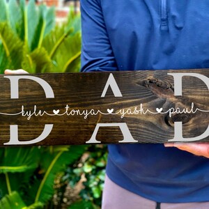 May include: A dark brown wooden sign with the word "DAD" in white letters. Below the word "DAD" is a line of text that reads "kyle &hearts; tonya &hearts; yash &hearts; paul".