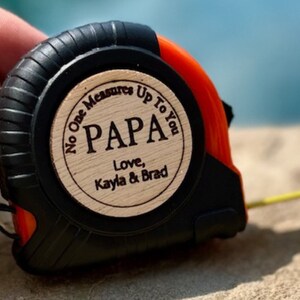 May include: A black and orange tape measure with a wooden circle attached to the front. The circle has the text "No One Measures Up To You PAPA Love, Kayla & Brad" engraved on it.