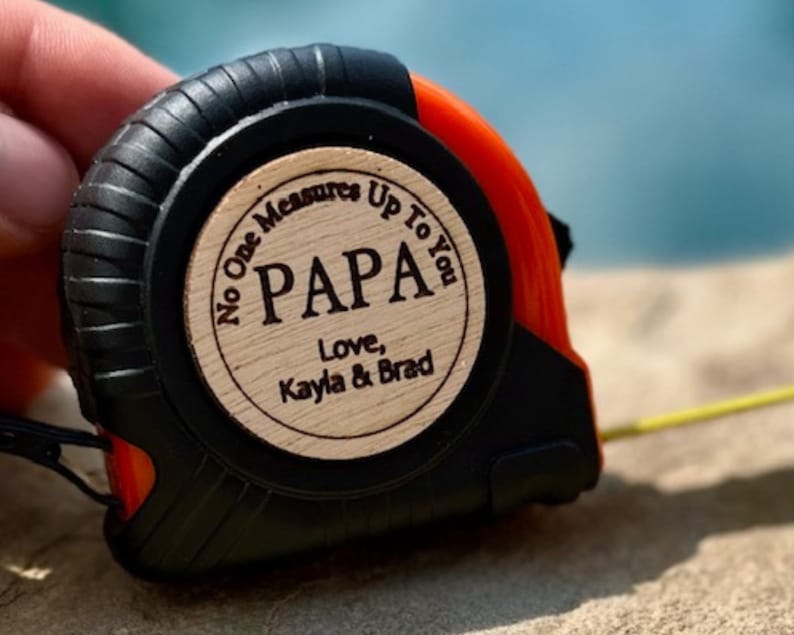 May include: A black and orange tape measure with a wooden circle attached to the front. The circle has the text "No One Measures Up To You PAPA Love, Kayla & Brad" engraved on it.